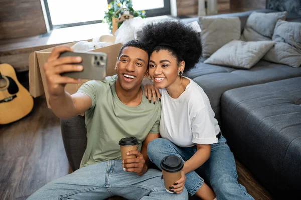Улыбающаяся африканская американская пара с кофе делает селфи на смартфоне рядом с коробками в новом доме — стоковое фото
