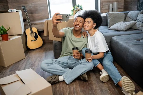Alegre casal afro-americano tomando selfie e segurando café perto de caixas de papelão em nova casa — Fotografia de Stock