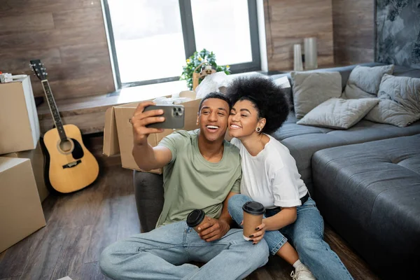 Feliz casal afro-americano tomando selfie e segurando café perto de pacotes na nova sala de estar — Fotografia de Stock