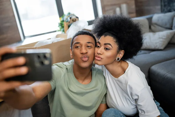 African american couple pouting lips while taking selfie on smartphone in new house — Stock Photo