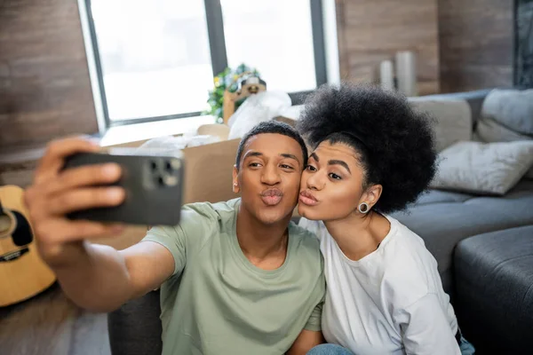 Casal afro-americano amamentando lábios enquanto toma selfie no smartphone na nova sala de estar — Fotografia de Stock