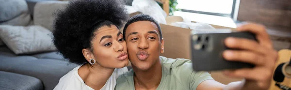 Couple afro-américain prendre selfie sur smartphone et bouder les lèvres dans une nouvelle maison, bannière — Photo de stock