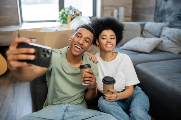 Alegre casal afro-americano tomando selfie e segurando café para ir perto de caixas em casa nova — Fotografia de Stock
