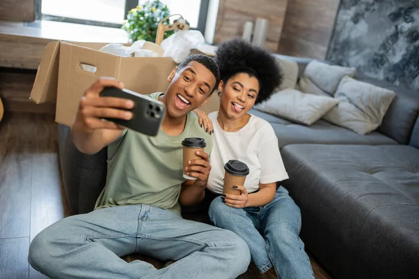 Excited african american couple taking selfie and holding coffee in living room in new house — Stock Photo
