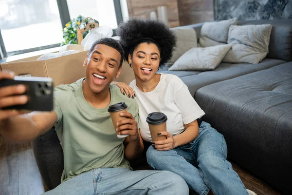 Pareja afroamericana haciendo muecas mientras toma selfie en el teléfono inteligente y la celebración de café en una casa nueva - foto de stock