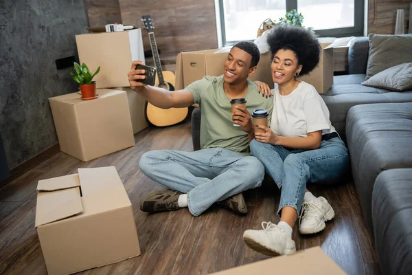 Pareja afroamericana haciendo muecas y tomando selfie con café cerca de cajas en casa nueva - foto de stock
