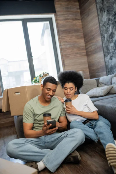 Pareja afro-americana positiva con café usando smartphone cerca de paquetes en casa nueva - foto de stock