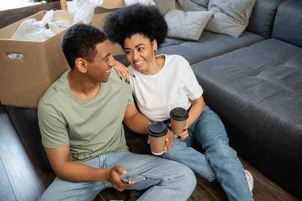 Positive african american couple with takeaway coffee using smartphone on floor in new house — Stock Photo