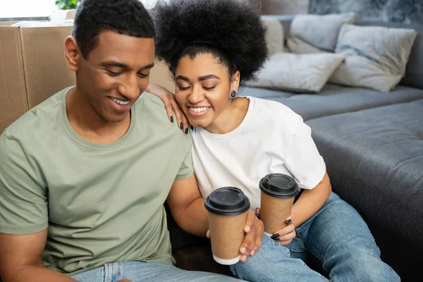 Gioiosa coppia afro-americana con caffè da asporto seduto vicino alla scatola di cartone nella nuova casa — Foto stock