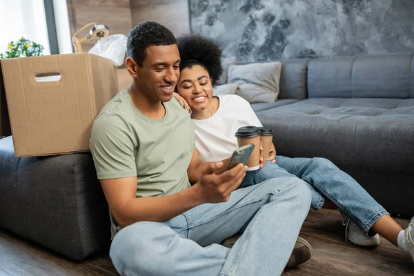 Positivo uomo afroamericano utilizzando smartphone vicino fidanzata con caffè per andare in una nuova casa — Foto stock