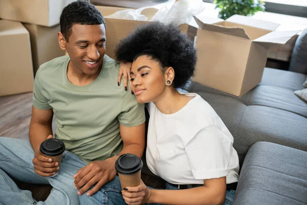 Pleased african american woman holding coffee to go near boyfriend and packages in new home — Stock Photo