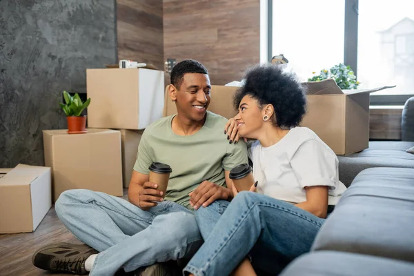 Donna afroamericana sorridente al fidanzato con caffè vicino a confezioni di cartone in una nuova casa — Foto stock