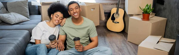 Pareja afroamericana complacida con café para llevar descansando cerca de cajas de cartón en casa nueva, pancarta - foto de stock