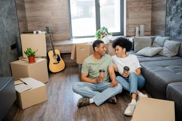Sonriente pareja afroamericana hablando y sosteniendo café cerca de cajas de cartón en nueva sala de estar - foto de stock