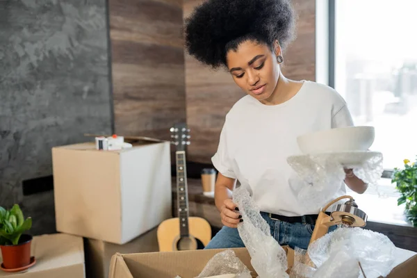 Mulher afro-americana desempacotando utensílios de mesa perto de caixa de papelão em nova casa — Fotografia de Stock