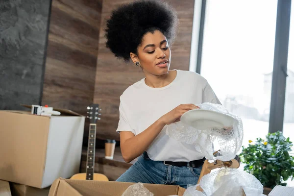 Femme afro-américaine déballage vaisselle près des emballages en carton dans une nouvelle maison — Photo de stock