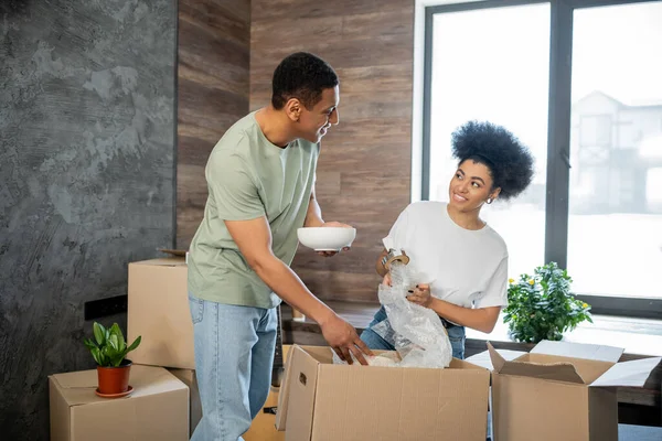 Sourire couple afro-américain parler tout en déballant la vaisselle près des boîtes dans une nouvelle maison — Photo de stock