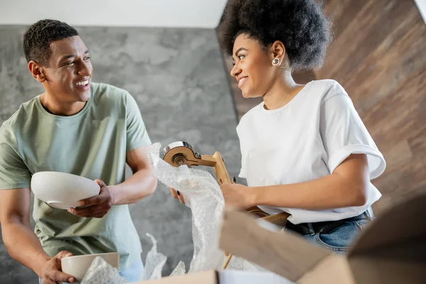 Cheerful african american couple unpacking tableware and lamp near carton box in new house — Stock Photo