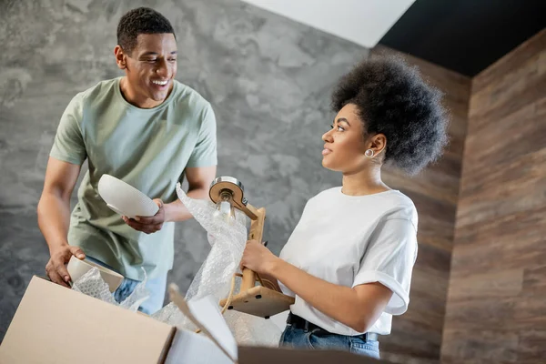 Fröhliche afrikanisch-amerikanische Mann auspackt Geschirr in der Nähe Freundin mit Lampe und Boxen in neuem Haus — Stockfoto