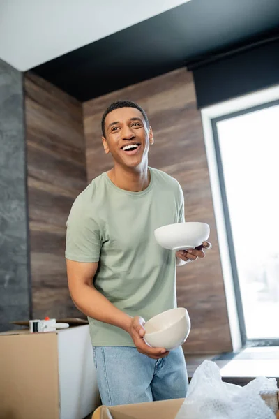 Cheerful african american man holding tableware near cardboard boxes in new house — Stock Photo