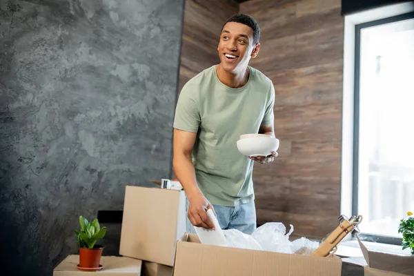 Hombre afroamericano positivo sosteniendo vajilla mientras desempaqueta cajas de cartón en casa nueva - foto de stock