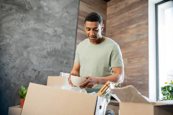 Joyful african american man holding plate while unpacking cardboard boxes in new house — Stock Photo