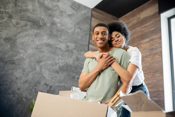 Smiling african american woman hugging boyfriend while standing near packages in new house — Stock Photo