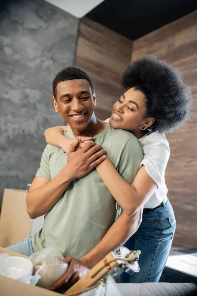Alegre afro-americana mulher abraçando namorado perto caixa de papelão na nova casa — Fotografia de Stock