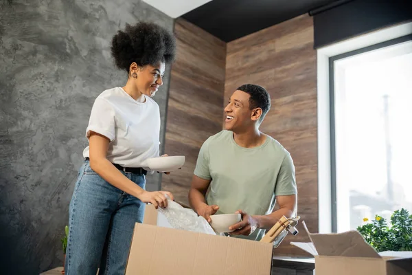 Улыбающаяся африканская американка держит тарелку и разговаривает с парнем распаковывая коробку в новом доме — стоковое фото