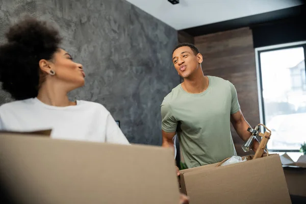 African american couple pouting lips while holding carton packages during moving in new house — Stock Photo