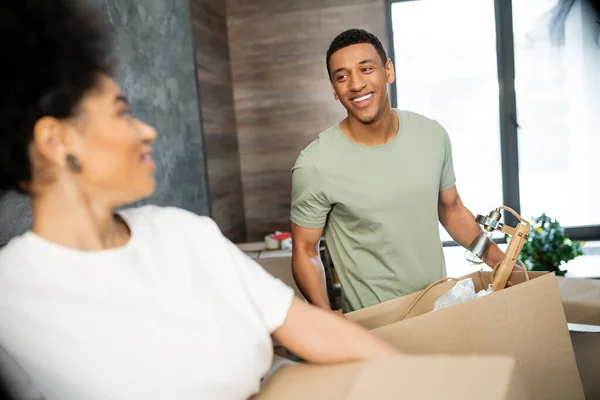 Carefree african american man holding cardboard box near blurred girlfriend in new house — Stock Photo