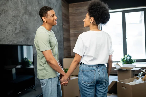 Überglückliches afrikanisch-amerikanisches Paar hält Händchen, während es neben Kartons in neuem Haus steht — Stockfoto