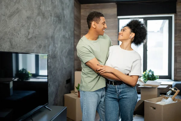 Heureux afro-américain homme étreignant petite amie près floue boîtes en carton dans la nouvelle maison — Photo de stock