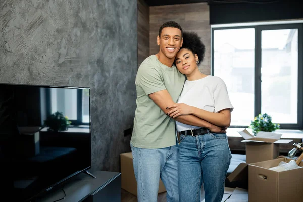 Sorridente africano americano uomo abbracciare fidanzata e guardando la fotocamera vicino scatole in nuova casa — Foto stock