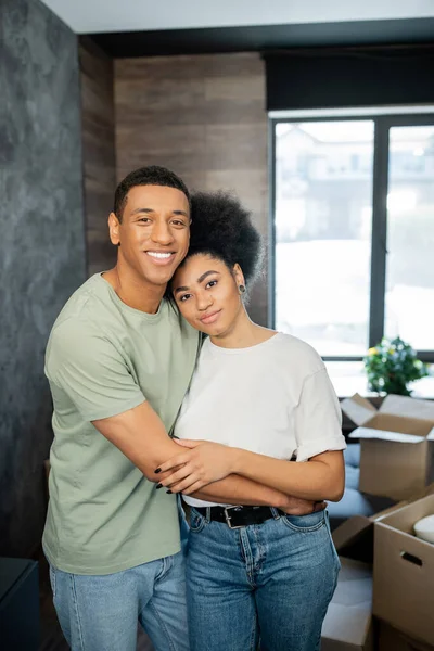 Ritratto di sorridente coppia afro-americana che abbraccia vicino scatole di cartone sfocate in una nuova casa — Foto stock
