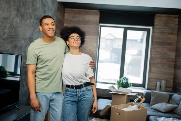 Verträumtes und lächelndes afrikanisch-amerikanisches Paar umarmt sich in der Nähe von Caron-Boxen im neuen Haus — Stockfoto