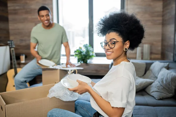 Smiling african american woman holding tableware near carton boxes and blurred boyfriend, new house — Stock Photo