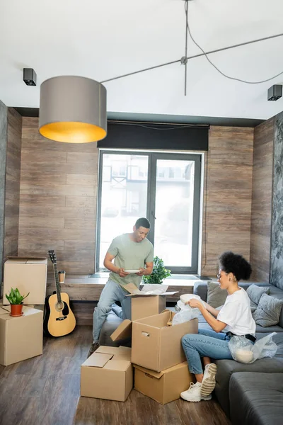 Positive african american couple holding tableware near cardboard boxes in new living room — Stock Photo