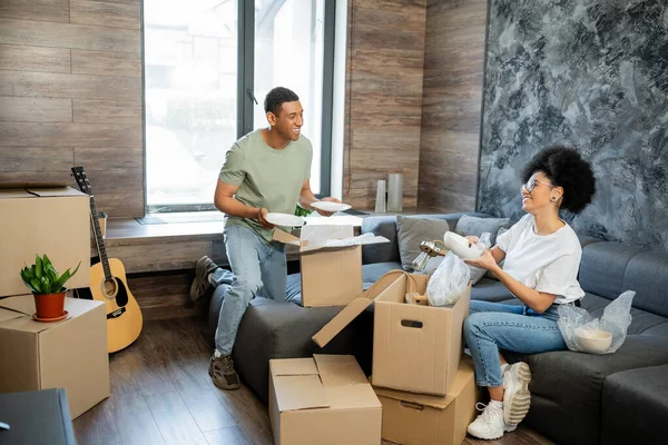 Alegre casal afro-americano desempacotando pacotes de papelão em nova sala de estar em casa — Fotografia de Stock