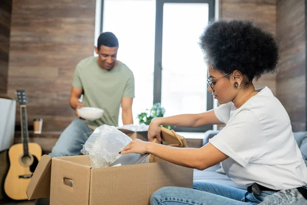 Africano americano mulher desembalagem caixa de papelão perto namorado borrado durante a mudança na nova casa — Fotografia de Stock