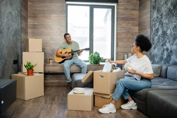 Sorridente afro-americano homem tocando guitarra acústica perto de namorada e caixas de papelão em nova casa — Fotografia de Stock