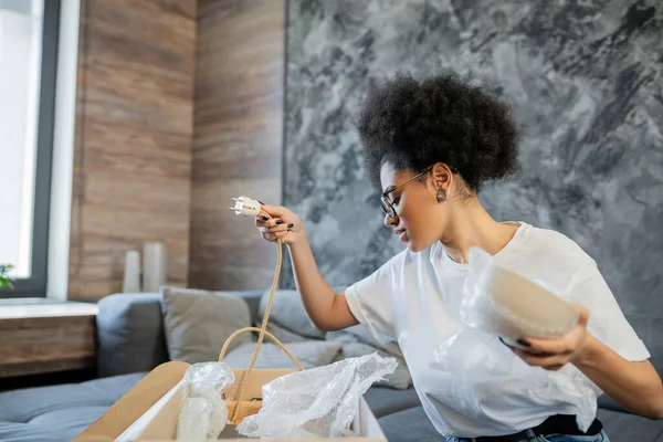 Vista lateral da mulher afro-americana segurando placa e arame enquanto desempacotando caixa na nova casa — Fotografia de Stock