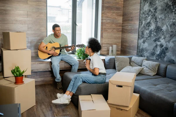 Alegre pareja afroamericana con café tocando guitarra acústica cerca de cajas en casa nueva - foto de stock