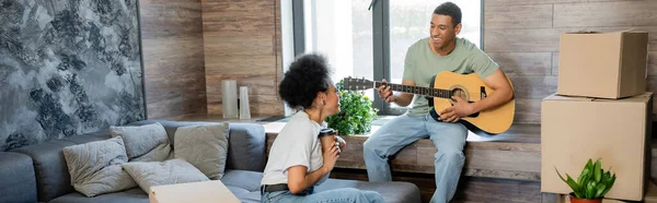 Feliz pareja afroamericana con café tocando la guitarra acústica cerca de paquetes en casa nueva, pancarta - foto de stock