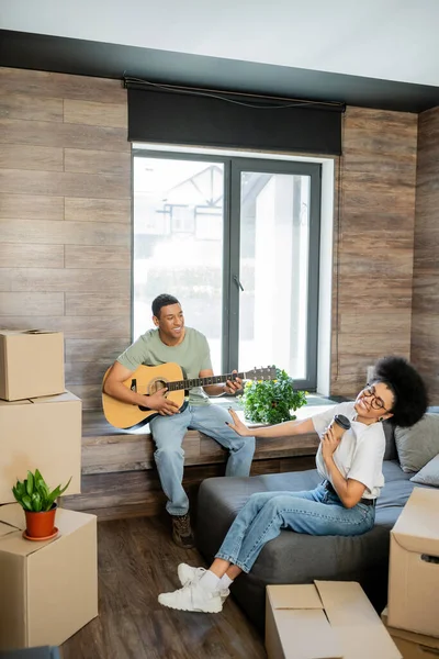 Щаслива афроамериканка тримає каву біля хлопця, граючи на акустичній гітарі в новому будинку — стокове фото