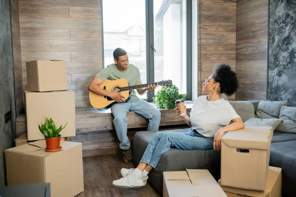Heureux homme afro-américain jouer de la guitare acoustique près de petite amie avec café à emporter dans une nouvelle maison — Photo de stock