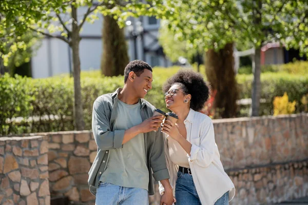 Joyeux couple afro-américain tenant la main et café pour aller tout en marchant dans la rue urbaine — Photo de stock