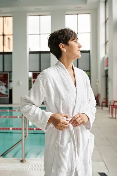Middle aged woman with short hair standing in white robe near swimming pool, sport, healthy life — Stock Photo