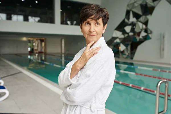 Mulher de meia-idade satisfeito com cabelo curto em pé em roupão branco, piscina, esporte, sorriso — Fotografia de Stock