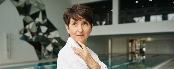 Middle aged woman with short hair standing in white robe, spa center, indoors swimming pool, banner — Stock Photo